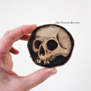 K&ouml;nnte beinhalten: Ein mit einem Brennstift gebranntes Sch&auml;delmotiv auf einer runden Holzscheibe. Der Sch&auml;del ist schwarz-wei&szlig; und hat ein detailliertes Design. Der Text "Agni Prasada Burning" steht oben auf der Scheibe.