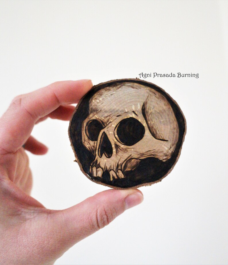 K&ouml;nnte beinhalten: Ein mit einem Brennstift gebranntes Sch&auml;delmotiv auf einer runden Holzscheibe. Der Sch&auml;del ist schwarz-wei&szlig; und hat ein detailliertes Design. Der Text "Agni Prasada Burning" steht oben auf der Scheibe.