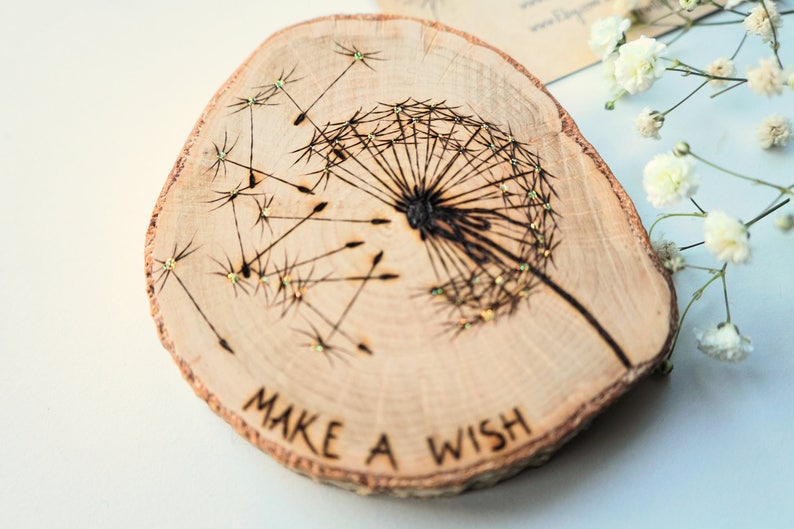 May include: A wooden round slice with a black and white dandelion design burned into the wood. The dandelion has gold glitter accents. The words "Make a Wish" are written below the dandelion.
