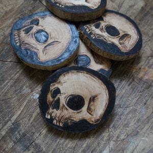 K&ouml;nnte beinhalten: Satz mit vier Untersetzern aus Holz mit Totenkopf-Design. Jeder Untersetzer besteht aus einer nat&uuml;rlichen Holzscheibe und hat ein schwarzes und wei&szlig;es Totenkopf-Design, das in das Holz gebrannt ist.