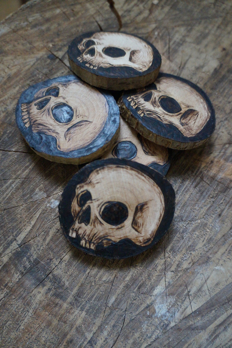 K&ouml;nnte beinhalten: Satz mit vier Untersetzern aus Holz mit Totenkopf-Design. Jeder Untersetzer besteht aus einer nat&uuml;rlichen Holzscheibe und hat ein schwarzes und wei&szlig;es Totenkopf-Design, das in das Holz gebrannt ist.