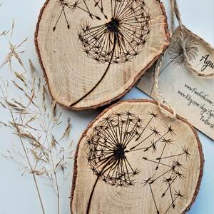 May include: Two wooden slices with a burnt design of a dandelion with seeds blowing in the wind. The wood is light brown with dark brown markings.