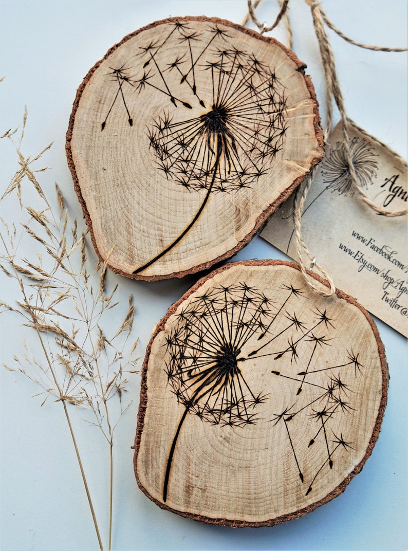 May include: Two wooden slices with a burnt design of a dandelion with seeds blowing in the wind. The wood is light brown with dark brown markings.