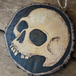 K&ouml;nnte beinhalten: Ein Holzscheiben-Anh&auml;nger mit einem schwarzen und wei&szlig;en Totenkopf-Design, das in das Holz gebrannt ist. Die Holzscheibe ist rund und hat einen nat&uuml;rlichen Rindenrand.