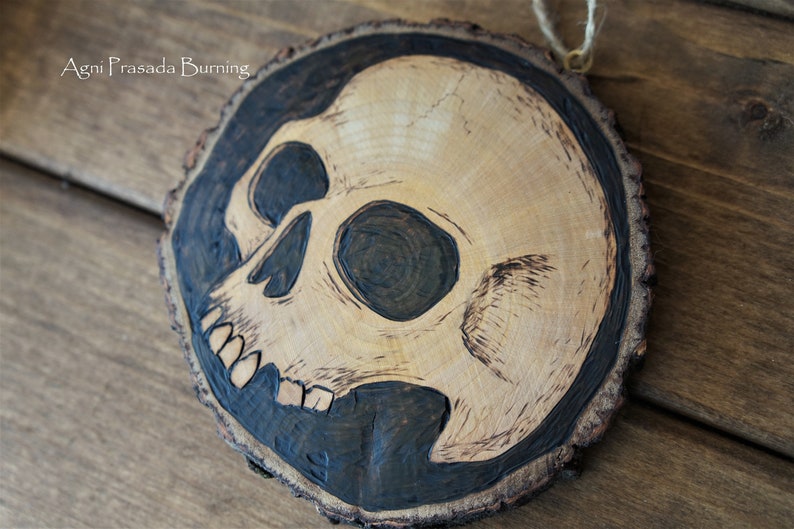 K&ouml;nnte beinhalten: Ein Holzscheiben-Anh&auml;nger mit einem schwarzen und wei&szlig;en Totenkopf-Design, das in das Holz gebrannt ist. Die Holzscheibe ist rund und hat einen nat&uuml;rlichen Rindenrand.