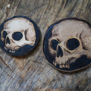 K&ouml;nnte beinhalten: Zwei Untersetzer aus Holz mit einem eingebrannten Sch&auml;delmotiv. Die Sch&auml;del sind wei&szlig; mit schwarzen Augen und auf einem dunkelbraunen Hintergrund.