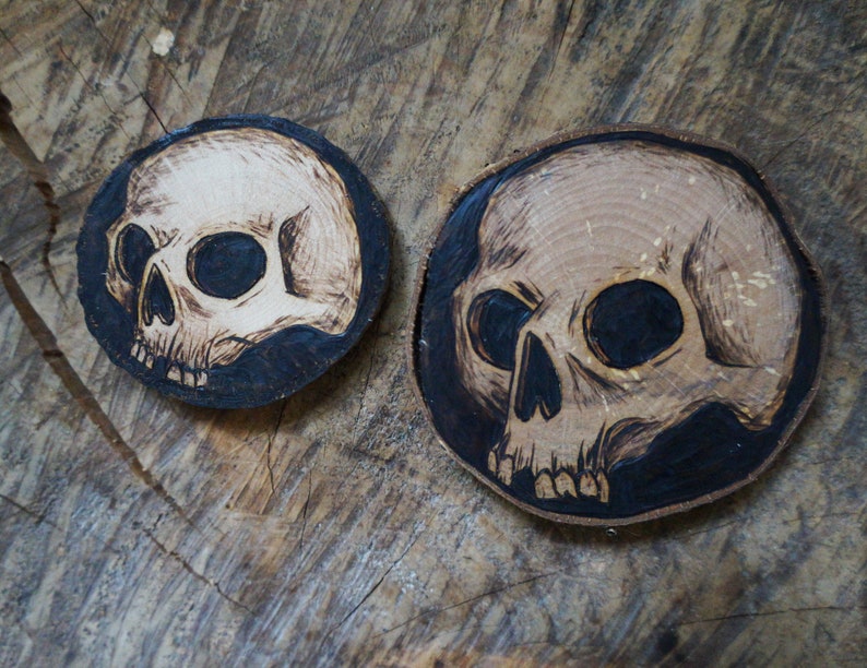 K&ouml;nnte beinhalten: Zwei Untersetzer aus Holz mit einem eingebrannten Sch&auml;delmotiv. Die Sch&auml;del sind wei&szlig; mit schwarzen Augen und auf einem dunkelbraunen Hintergrund.
