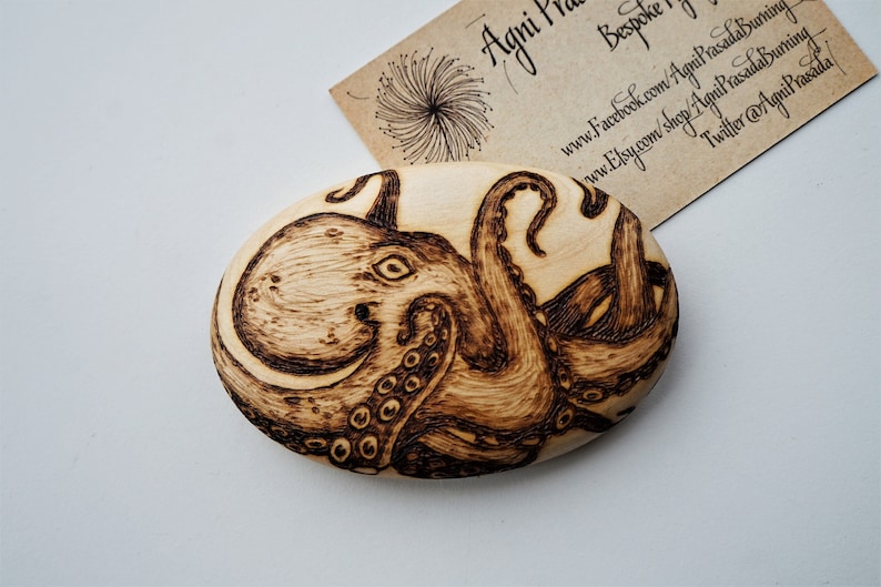 May include: A woodburned design of an octopus on a wooden oval. The octopus is brown and black and has eight tentacles.