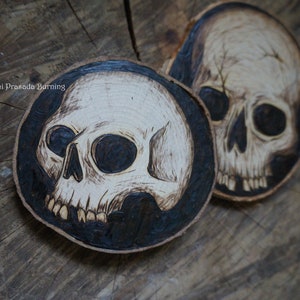 K&ouml;nnte beinhalten: Zwei Untersetzer aus Holz mit einem eingebrannten Sch&auml;delmotiv. Die Sch&auml;del sind wei&szlig; mit schwarzen Augen und auf einem schwarzen Hintergrund.