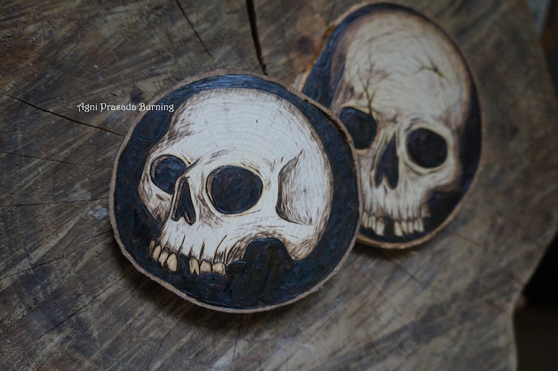 K&ouml;nnte beinhalten: Zwei Untersetzer aus Holz mit einem eingebrannten Sch&auml;delmotiv. Die Sch&auml;del sind wei&szlig; mit schwarzen Augen und auf einem schwarzen Hintergrund.