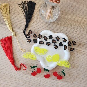 Peut inclure: Plusieurs éléments décoratifs. Trois pompons dorés, noirs et rouges. Un plat blanc en forme de nuage avec des motifs léopard et des tranches de citron. Une feuille transparente avec des graphiques de cerises.