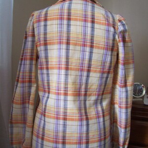 May include: A long-sleeved plaid shirt with a light yellow, orange, purple, and brown plaid pattern. The shirt has a button-up collar and long sleeves with elastic cuffs.
