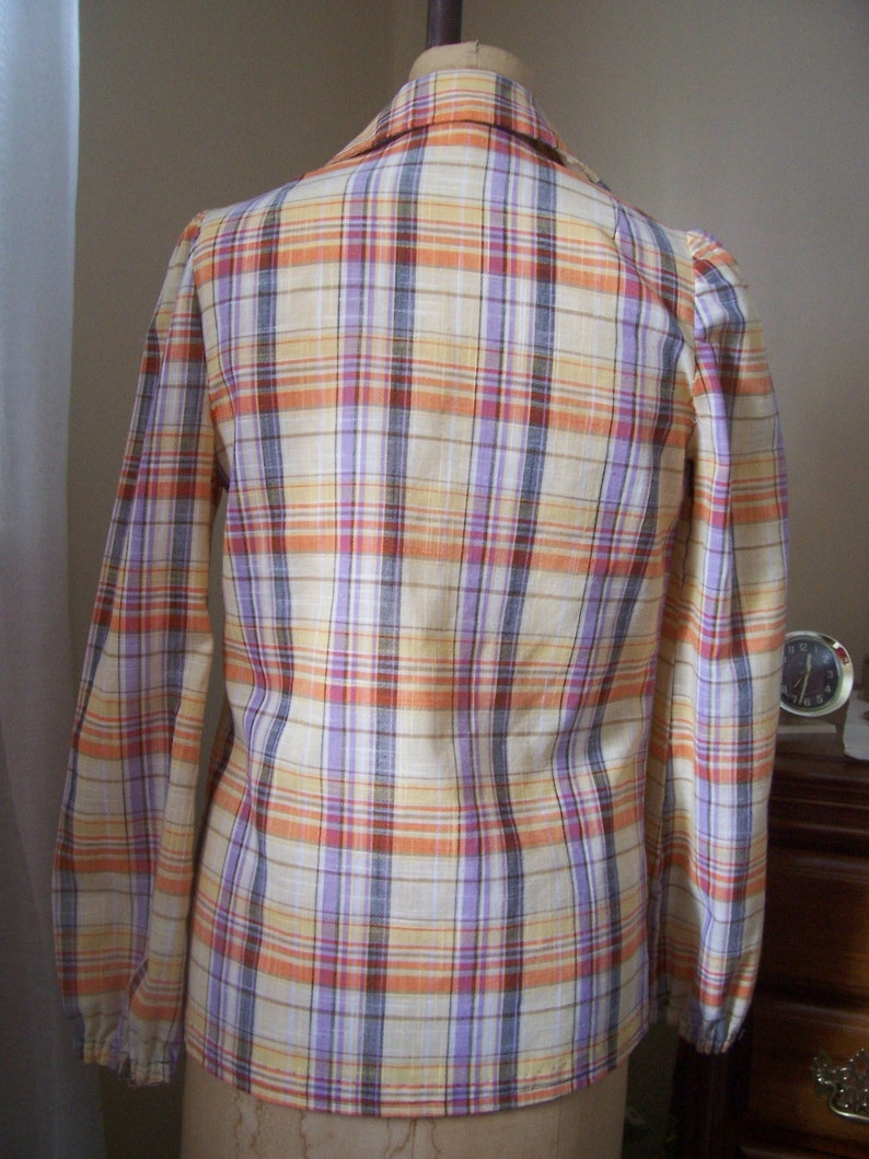 May include: A long-sleeved plaid shirt with a light yellow, orange, purple, and brown plaid pattern. The shirt has a button-up collar and long sleeves with elastic cuffs.