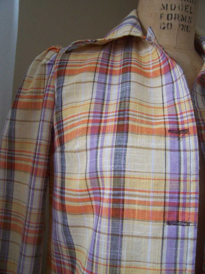 May include: A plaid shirt with a light yellow, orange, purple, and brown plaid pattern. The shirt has a button-up collar and long sleeves.
