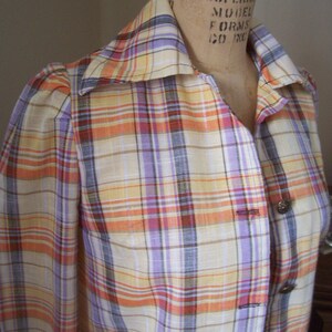 May include: A plaid shirt with a collared neckline and long sleeves. The shirt has a light yellow base with orange, purple, and brown stripes. The shirt has buttons down the front and is made of a lightweight fabric.