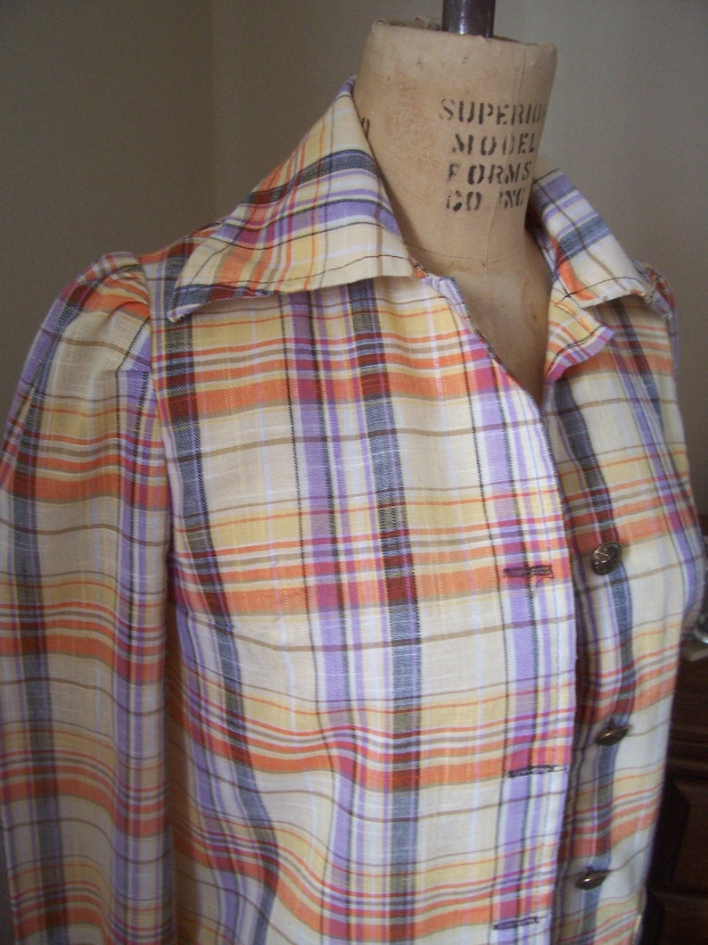 May include: A plaid shirt with a collared neckline and long sleeves. The shirt has a light yellow base with orange, purple, and brown stripes. The shirt has buttons down the front and is made of a lightweight fabric.