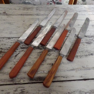 May include: A set of eight silver butter knives with brown plastic handles. The knives are arranged on a white wooden surface.