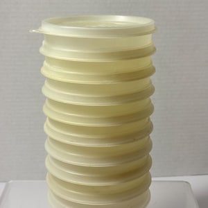 May include: A stack of ten circular, semi-translucent, off-white plastic lids. Each lid features a slightly raised rim and a small tab on the side for easy removal. The lids are uniform in size and shape, stacked vertically to form a cylindrical column.