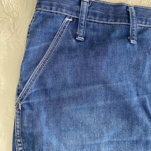 May include: Close-up of blue denim jeans. The jeans feature a classic design with a visible pocket and belt loops. White stitching accents the seams and pocket edges, creating a contrast against the blue fabric. The denim has a slightly faded appearance.