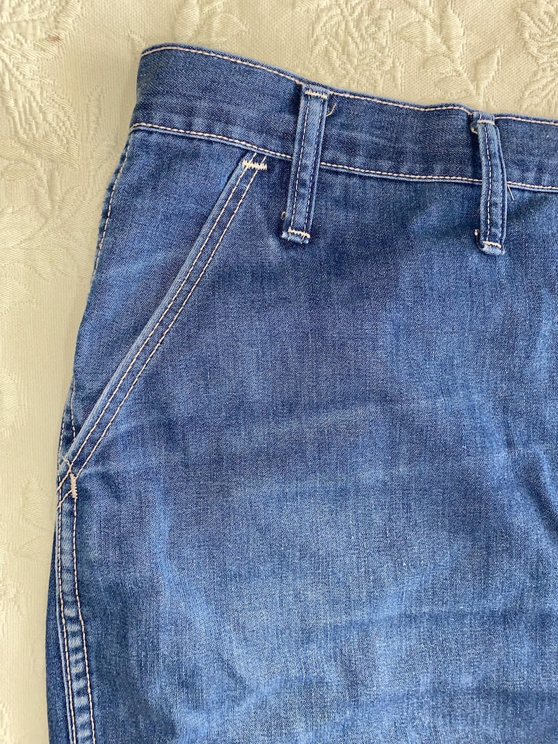 May include: Close-up of blue denim jeans. The jeans feature a classic design with a visible pocket and belt loops. White stitching accents the seams and pocket edges, creating a contrast against the blue fabric. The denim has a slightly faded appearance.