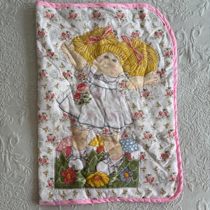 Op de afbeelding: Een gewatteerde babydeken met een poppenontwerp met blonde vlechten en een witte jurk. De deken heeft een bloemenpatroon en een roze rand. De pop staat op een grasveld met paddenstoelen en bloemen.