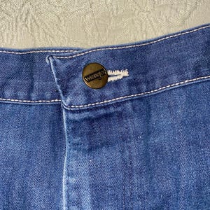 May include: Close-up of blue denim jeans with a button closure. The button is a brass color and has the word "Wrangler" embossed on it. White stitching is visible along the seams and around the button.