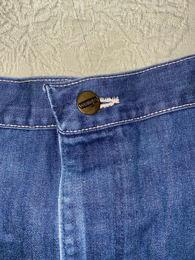 May include: Close-up of blue denim jeans with a button closure. The button is a brass color and has the word "Wrangler" embossed on it. White stitching is visible along the seams and around the button.