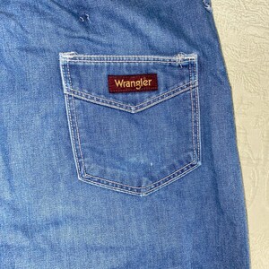 May include: Close-up of blue denim jeans with a back pocket. The pocket features white stitching and a rectangular brown label with the brand name "Wrangler" in white lettering. The jeans have a faded appearance.