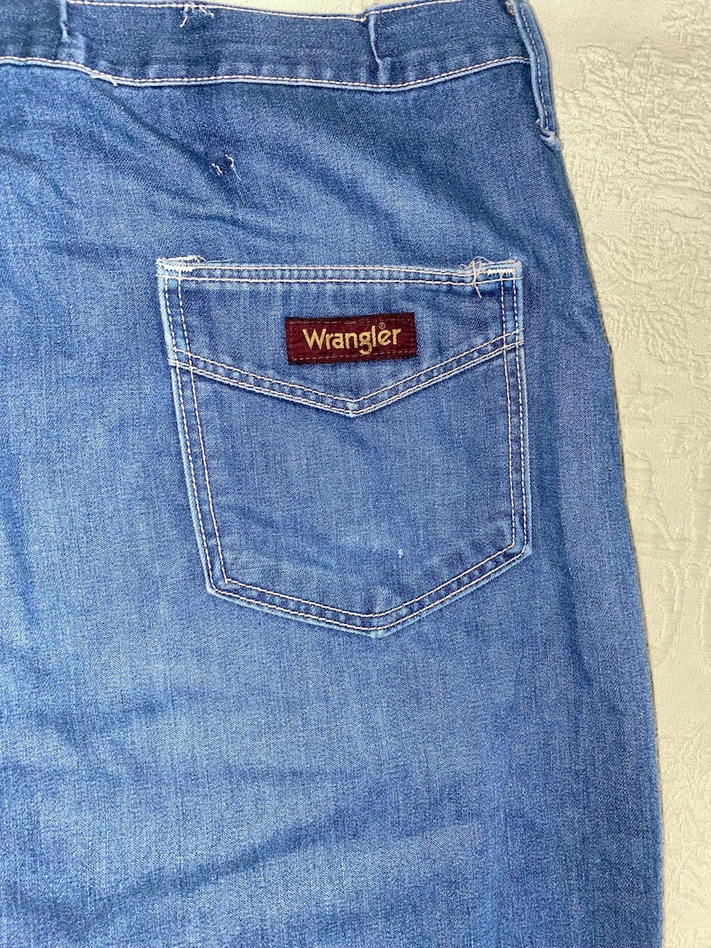 May include: Close-up of blue denim jeans with a back pocket. The pocket features white stitching and a rectangular brown label with the brand name "Wrangler" in white lettering. The jeans have a faded appearance.
