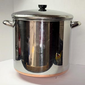 Op de afbeelding: Een grote, cilindrische soeppan van roestvrij staal met een gepolijste, reflecterende afwerking. De pan heeft twee zijhandgrepen, een bijpassend deksel met een zwarte knop en een opvallende koperkleurige bodem. Het metaal reflecteert de omgeving.