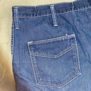 May include: Close-up of blue denim jeans with a back pocket. The jeans have white stitching and belt loops. The fabric appears to be a medium wash denim.