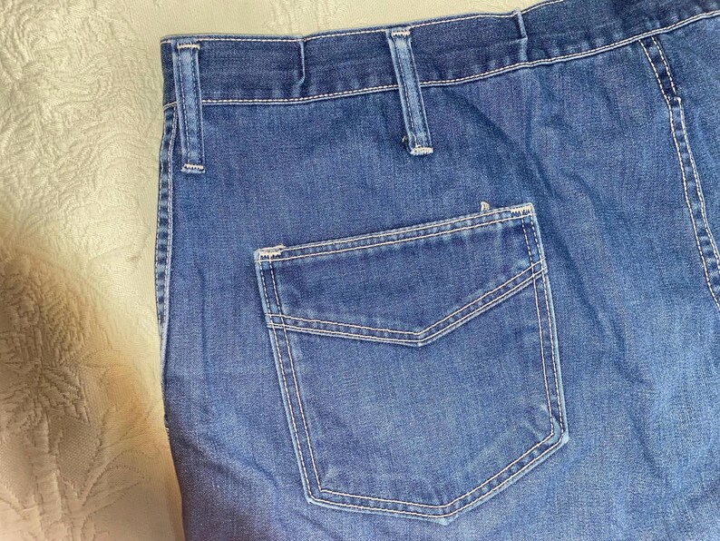May include: Close-up of blue denim jeans with a back pocket. The jeans have white stitching and belt loops. The fabric appears to be a medium wash denim.