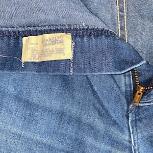 May include: Close-up of blue denim jeans with a zipper and a tag that reads "Wrangler Jeans For Men". The tag also includes washing instructions and the size "42 SHORT". The jeans have white stitching.