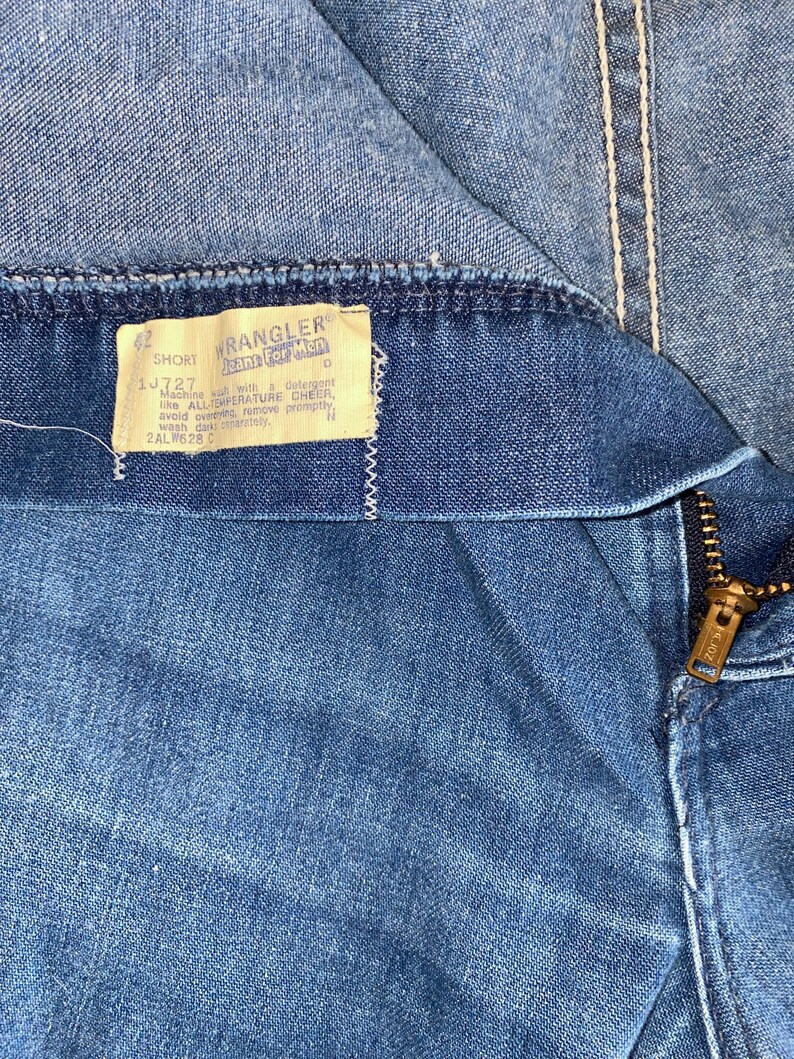 May include: Close-up of blue denim jeans with a zipper and a tag that reads "Wrangler Jeans For Men". The tag also includes washing instructions and the size "42 SHORT". The jeans have white stitching.