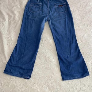 May include: Blue denim jeans with a flared leg design. The jeans have two back pockets and a small rectangular patch on the right back pocket. The jeans are laid flat on a white patterned surface.