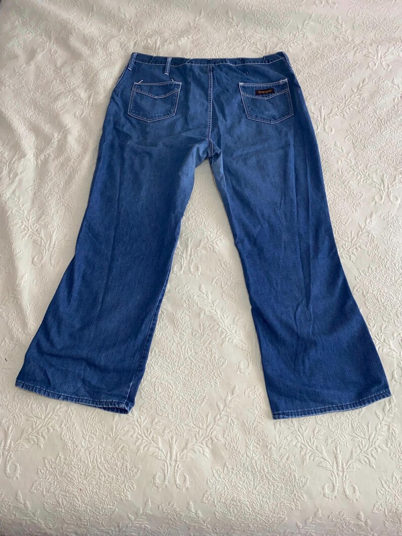 May include: Blue denim jeans with a flared leg design. The jeans have two back pockets and a small rectangular patch on the right back pocket. The jeans are laid flat on a white patterned surface.