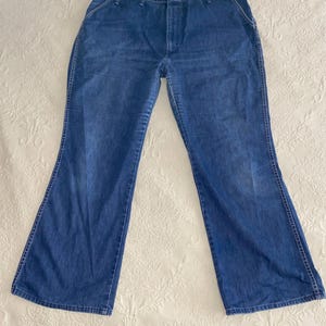 May include: Blue denim jeans with a flared leg design. The jeans have a classic five-pocket style, a button and zipper closure, and white stitching details. The jeans are laid flat on a white surface.