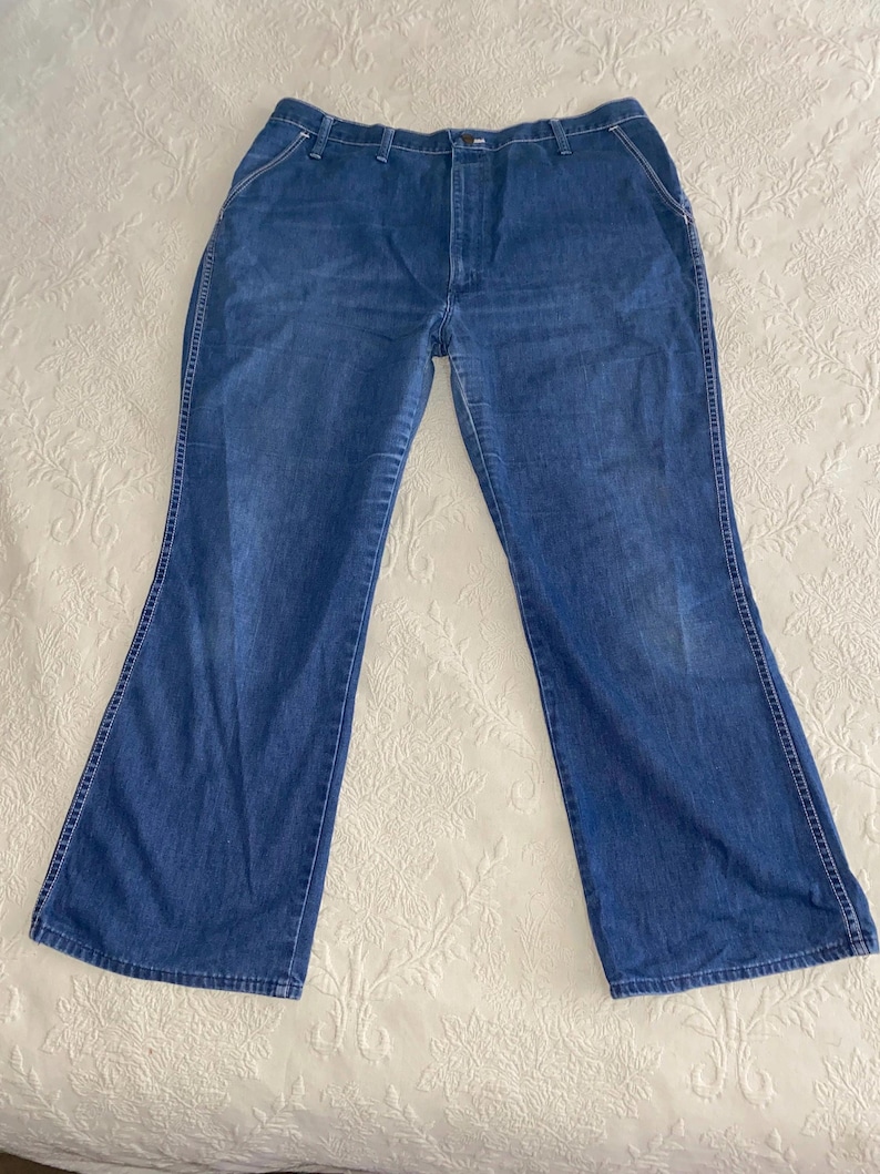 May include: Blue denim jeans with a flared leg design. The jeans have a classic five-pocket style, a button and zipper closure, and white stitching details. The jeans are laid flat on a white surface.