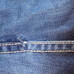 May include: Close-up of blue denim fabric with visible texture and white stitching. The stitching is a prominent design element, adding contrast to the denim. The image highlights the details of the textile.