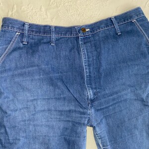 May include: Close-up of blue denim jeans. The jeans have a button closure, belt loops, and pockets. The stitching is white, and the denim has a faded appearance. The jeans are a classic style.