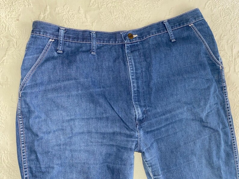 May include: Close-up of blue denim jeans. The jeans have a button closure, belt loops, and pockets. The stitching is white, and the denim has a faded appearance. The jeans are a classic style.