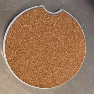 May include: A circular car coaster featuring a cork centre and a white edge. It has a small indentation on one side. The cork provides an absorbent surface. The coaster is designed to fit into a car's cup holder.