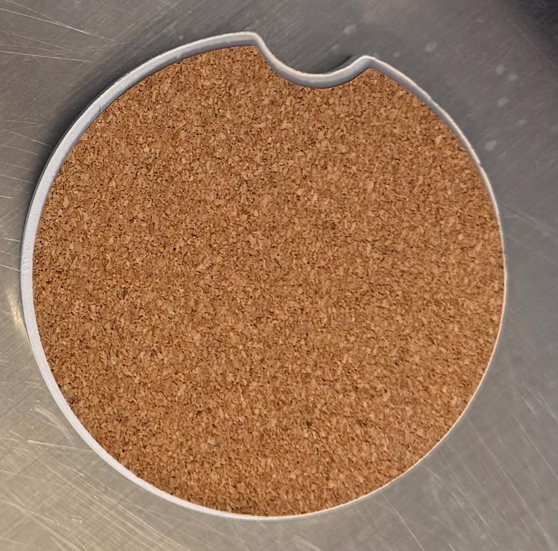 May include: A circular car coaster featuring a cork centre and a white edge. It has a small indentation on one side. The cork provides an absorbent surface. The coaster is designed to fit into a car's cup holder.
