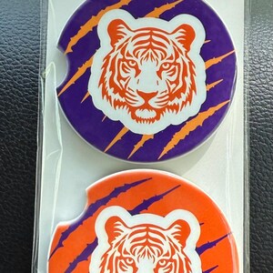 May include: Two car coasters with a tiger design. One coaster is purple with orange claw marks and the other is orange with purple claw marks. The tiger is white with orange details. The packaging reads "Car Coasters Handmade with love."