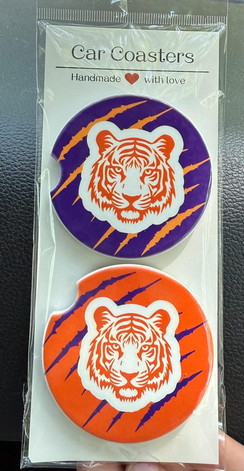 May include: Two car coasters with a tiger design. One coaster is purple with orange claw marks and the other is orange with purple claw marks. The tiger is white with orange details. The packaging reads "Car Coasters Handmade with love."