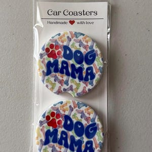 May include: Two car coasters with a colourful bone and paw print pattern. The coasters have the words "Dog Mama" in blue bubble letters. The packaging reads "Car Coasters" and "Handmade with love".