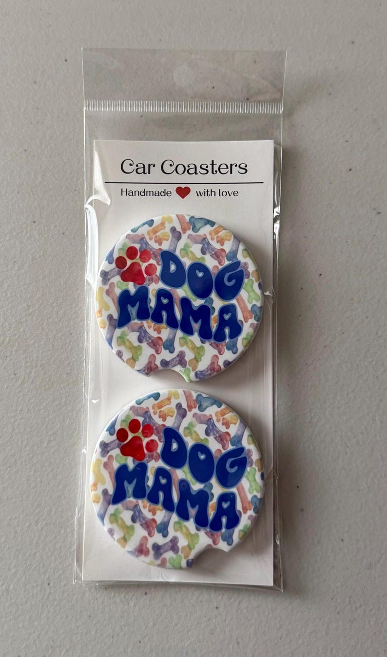 May include: Two car coasters with a colourful bone and paw print pattern. The coasters have the words "Dog Mama" in blue bubble letters. The packaging reads "Car Coasters" and "Handmade with love".