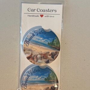 May include: Two car coasters with a beach scene design. The coasters feature a blue sky, ocean, sand, seashells, and palm trees. The words "Beach Vibes" are written on each coaster. The packaging reads "Car Coasters" and "Handmade with love."