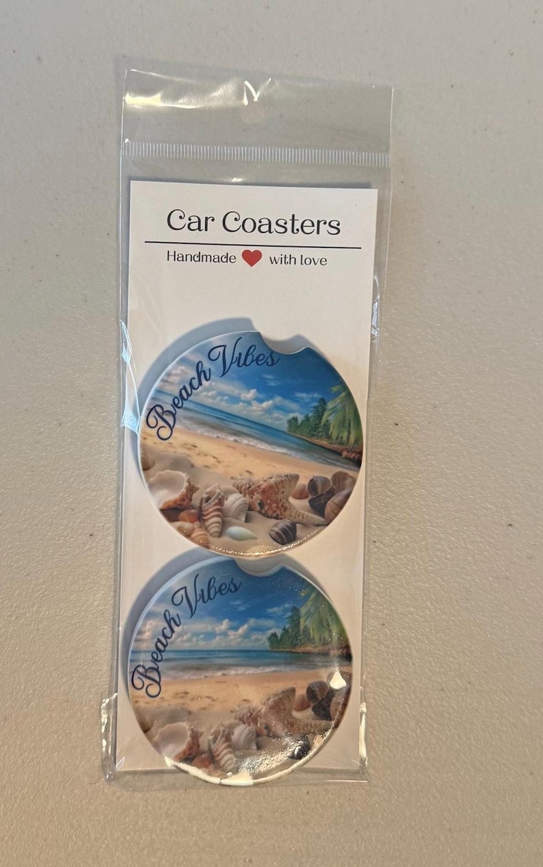 May include: Two car coasters with a beach scene design. The coasters feature a blue sky, ocean, sand, seashells, and palm trees. The words "Beach Vibes" are written on each coaster. The packaging reads "Car Coasters" and "Handmade with love."
