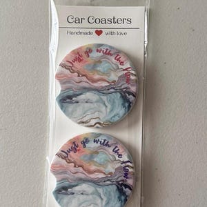 May include: Two car coasters with a colourful abstract design in pink, blue, and gold tones. The coasters have the text "Just go with the flow" in a cursive font. The packaging reads "Car Coasters, Handmade with love."
